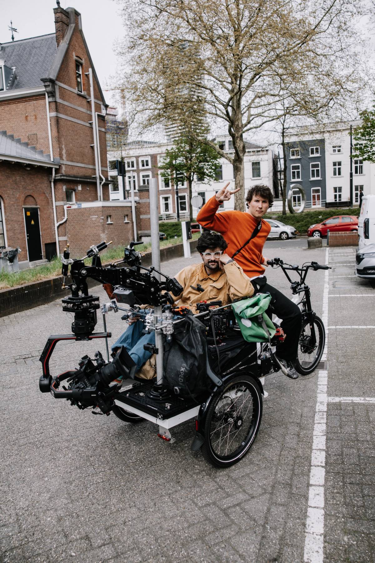Two crew members pose on a cargo bike loaded with cameras and production gear on a city street.