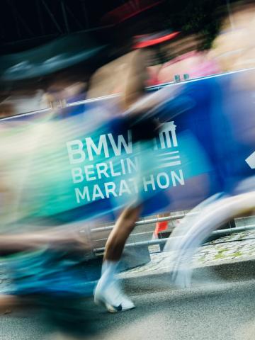 Bewegungsunscharfe Läufer passieren eine blaue Marathon-Absperrung am Straßenrand.