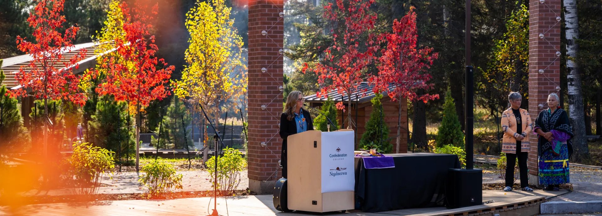 Speaker at a podium during an outdoor campus ceremony surrounded by autumn trees