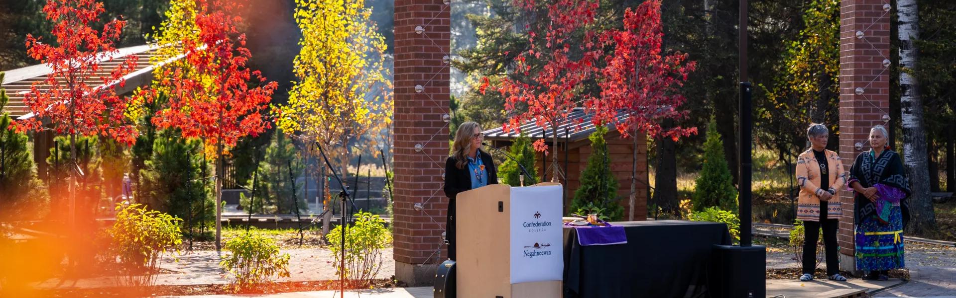 Speaker at a podium during an outdoor campus ceremony surrounded by autumn trees