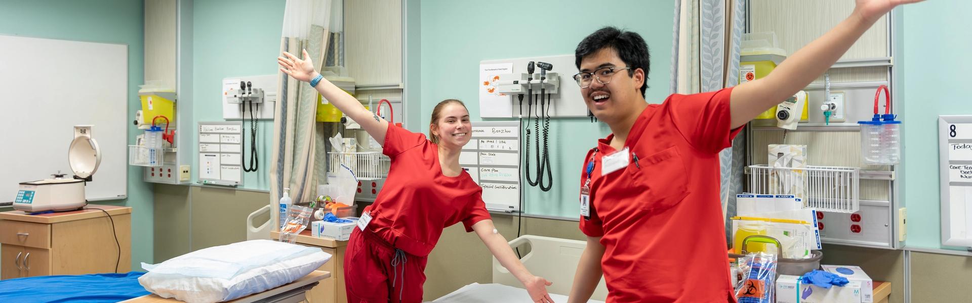 Two students in red medical scrubs stand in a simulated patient room with hospital beds, side tables, and wall-mounted medical equipment.