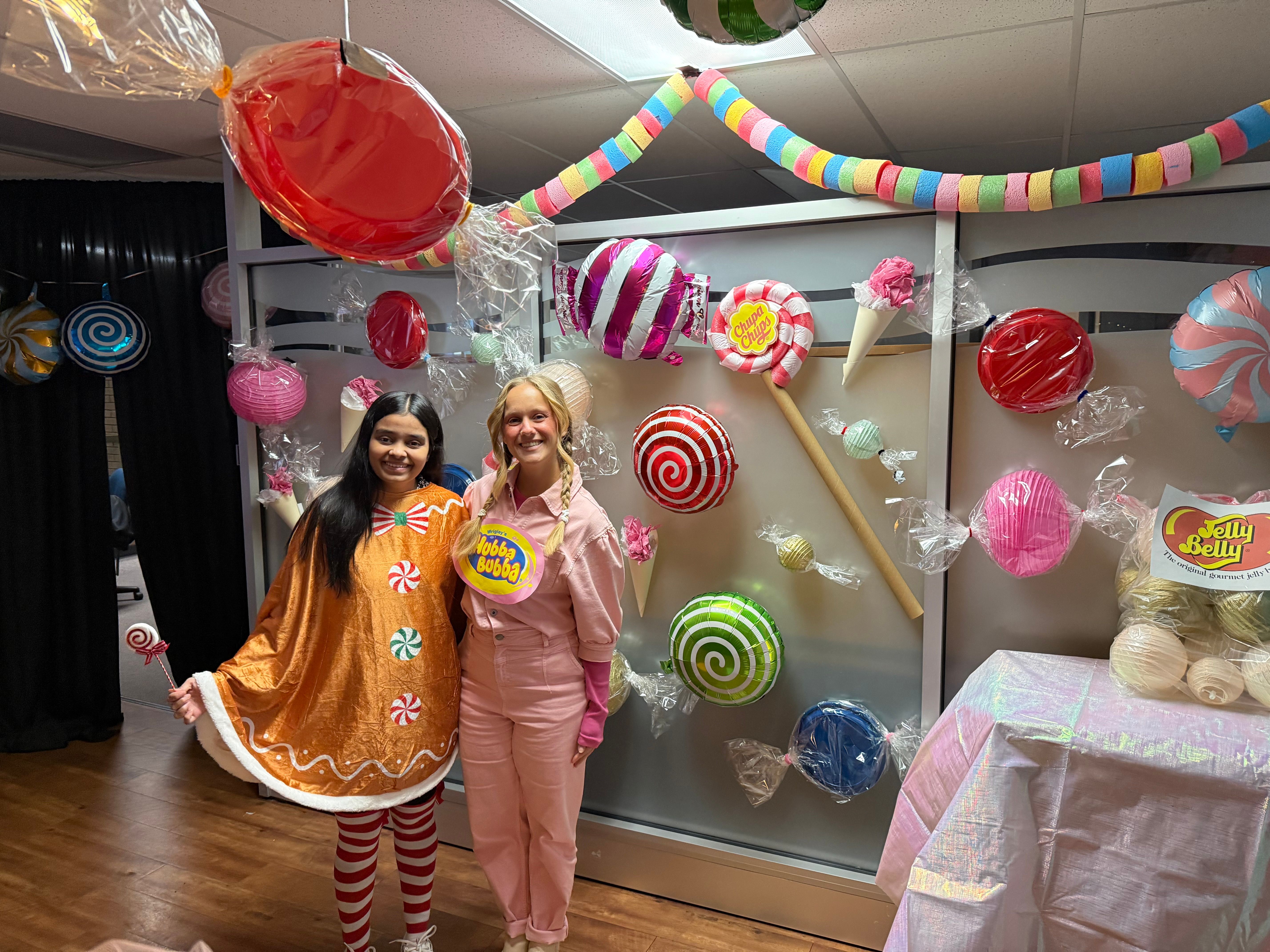 Two people in candy-themed costumes stand beside a Halloween display decorated with oversized lollipops and colourful paper garlands.