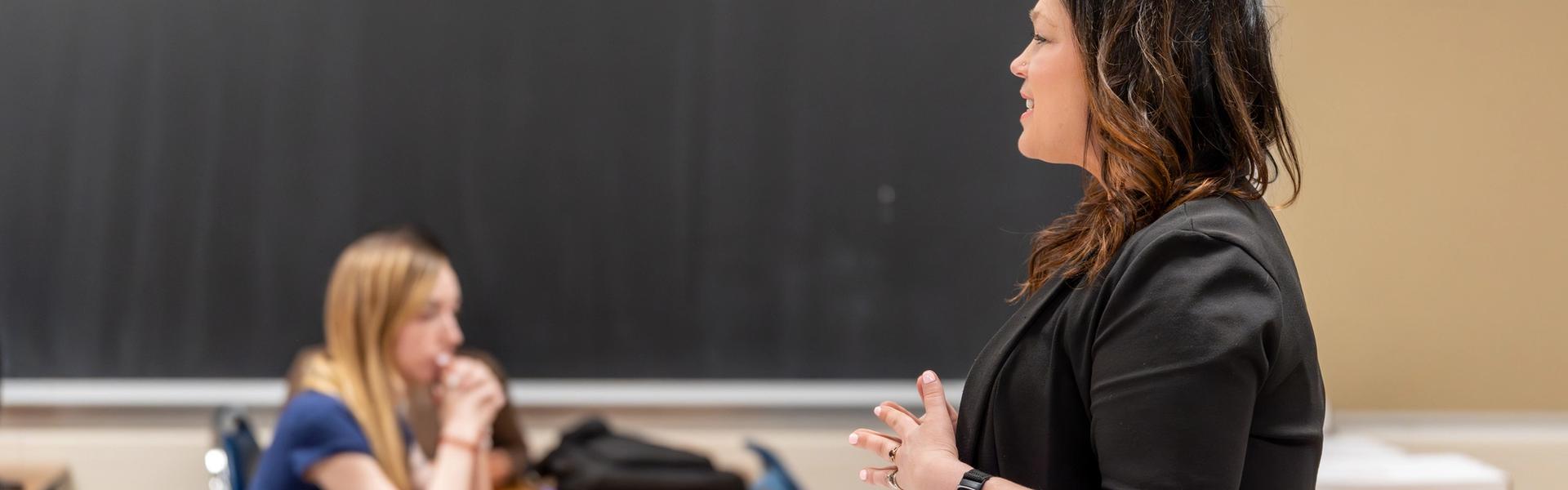 A woman in a black blazer speaks to a seated student in a classroom. The room has wooden desks, a large chalkboard, and a casual academic setting