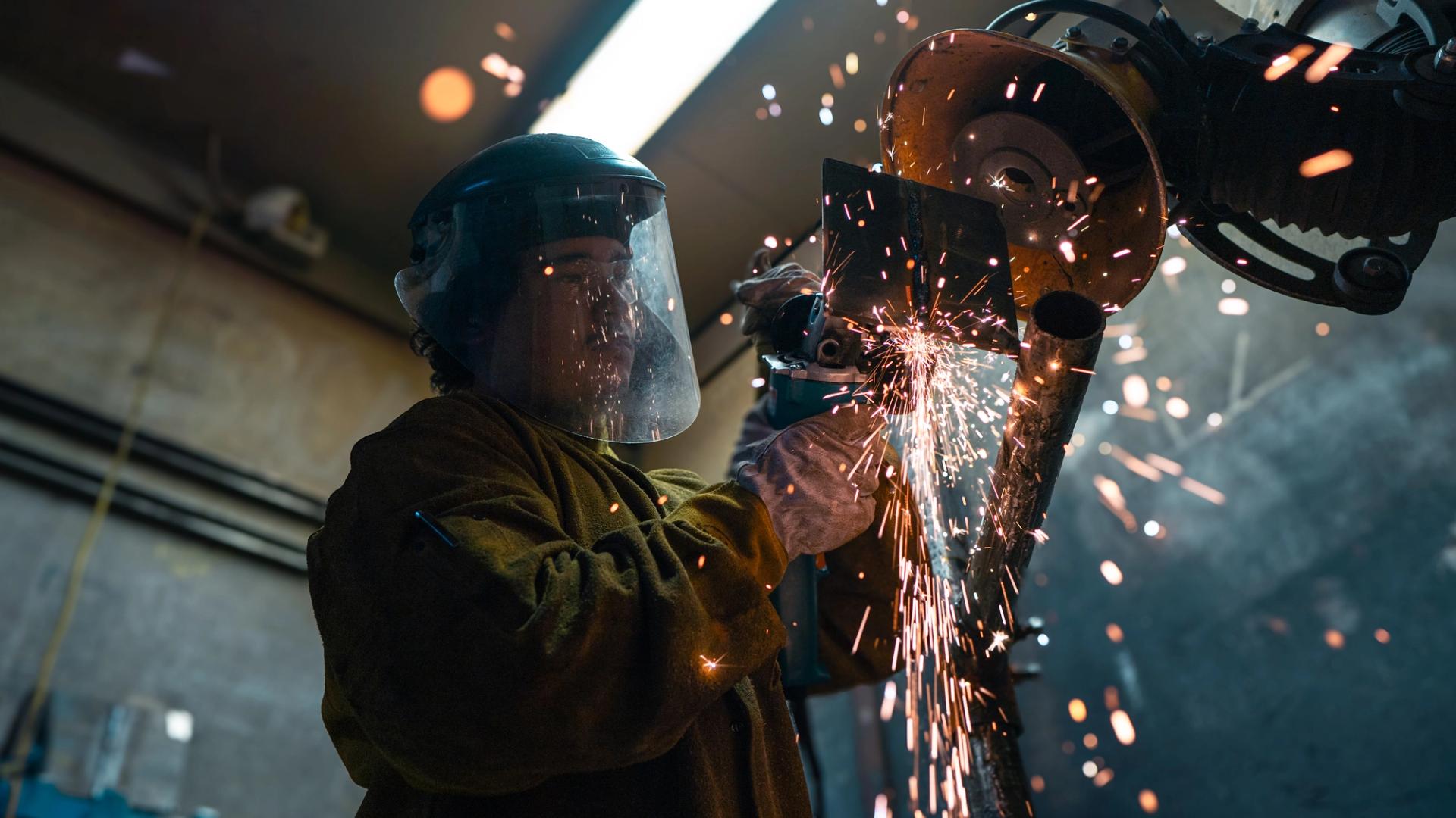 Student using a chop saw to cut metal pipe, wearing a face shield as sparks fly in a trades workshop.