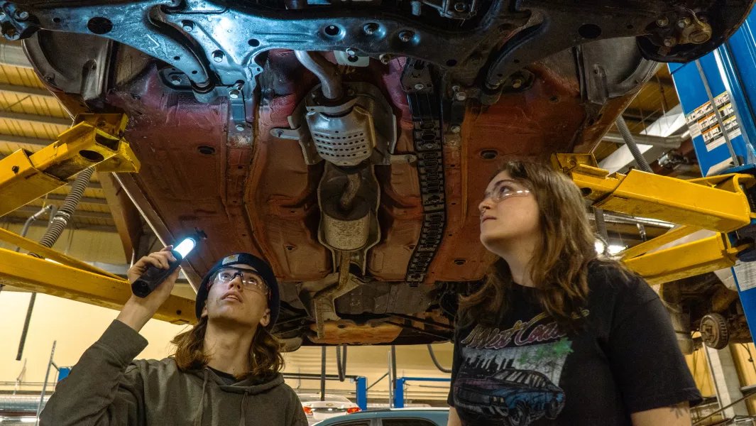 Two students inspecting the underside of a car with a flashlight in a workshop