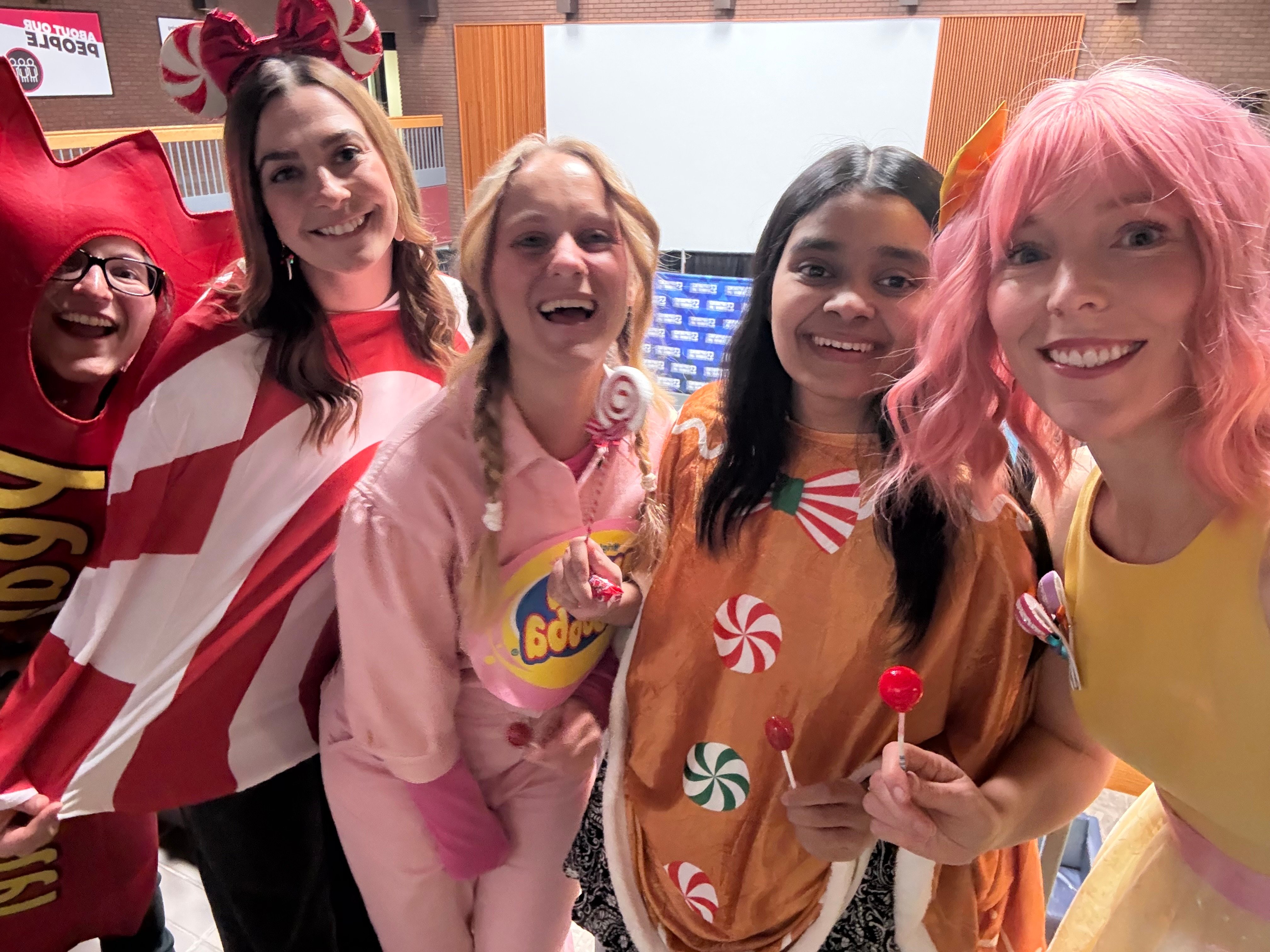 Five people pose in colourful Halloween costumes, including candy-themed outfits and a red-and-white striped cape, indoors.