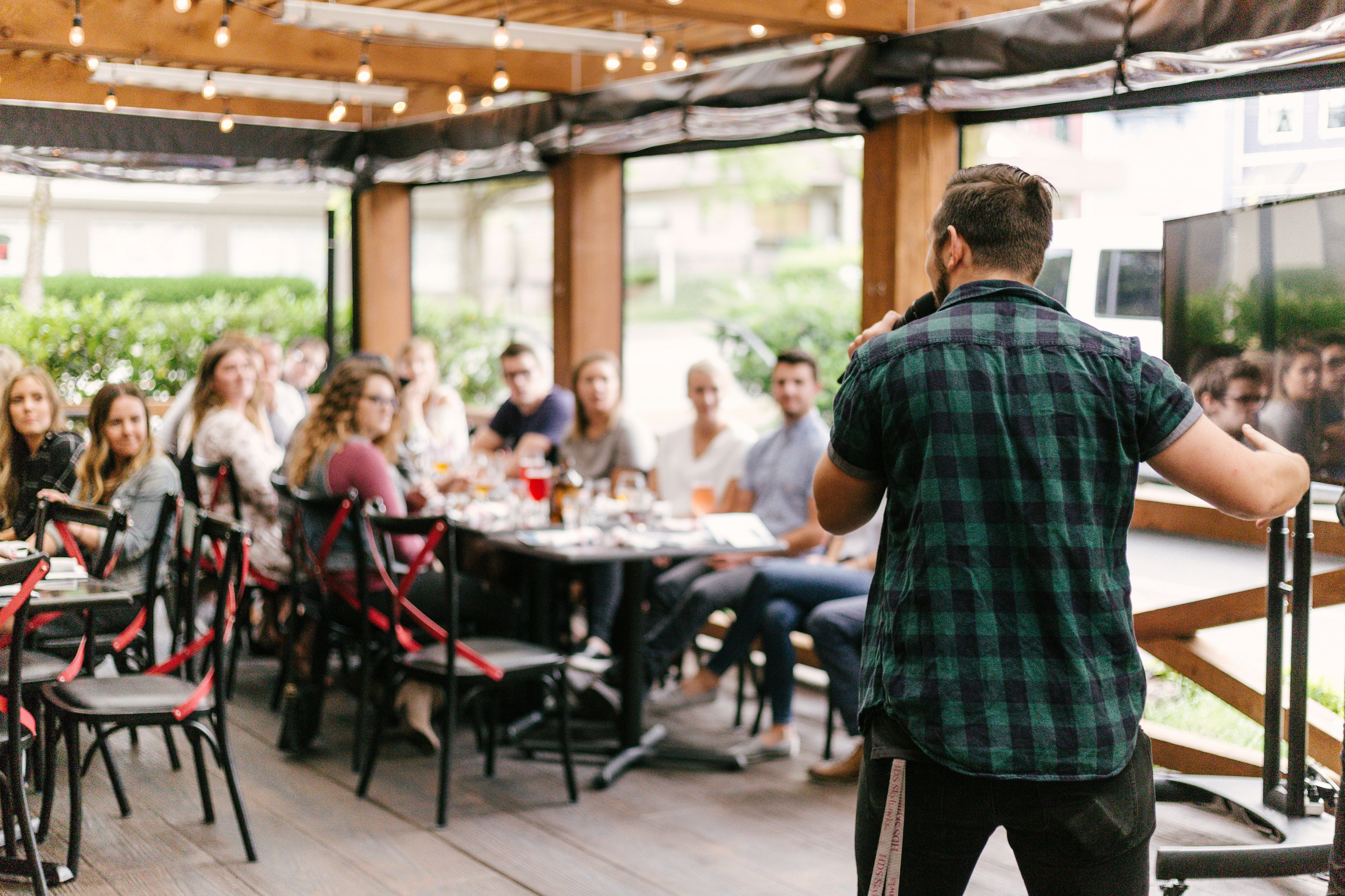 Man speaking to a seated group at tables on a covered patio