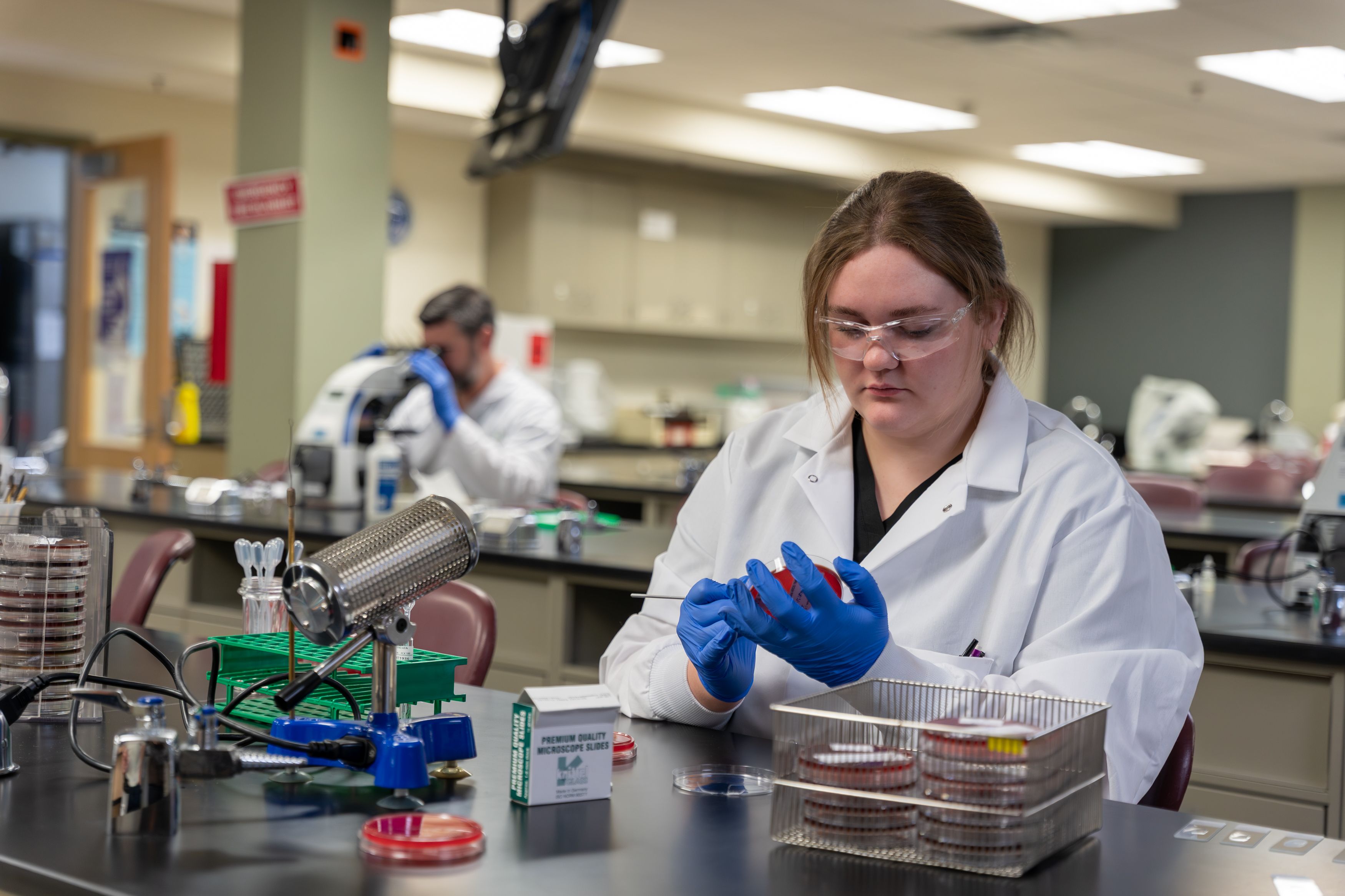 Student in lab coat preparing a petri dish sample at a laboratory workstation