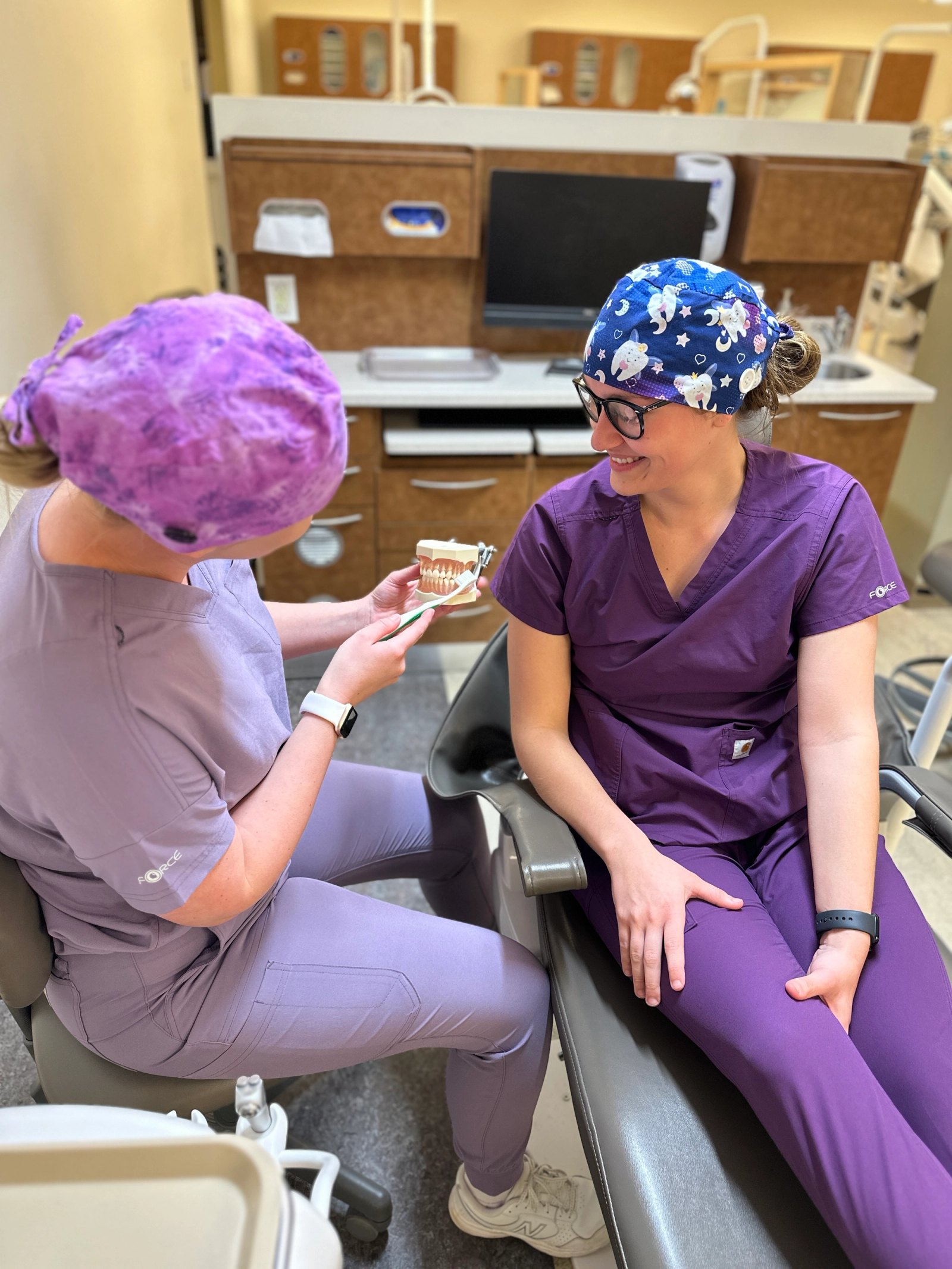 Two people in purple scrubs sit in a dental clinic, one holding a tooth model and toothbrush while smiling at the other