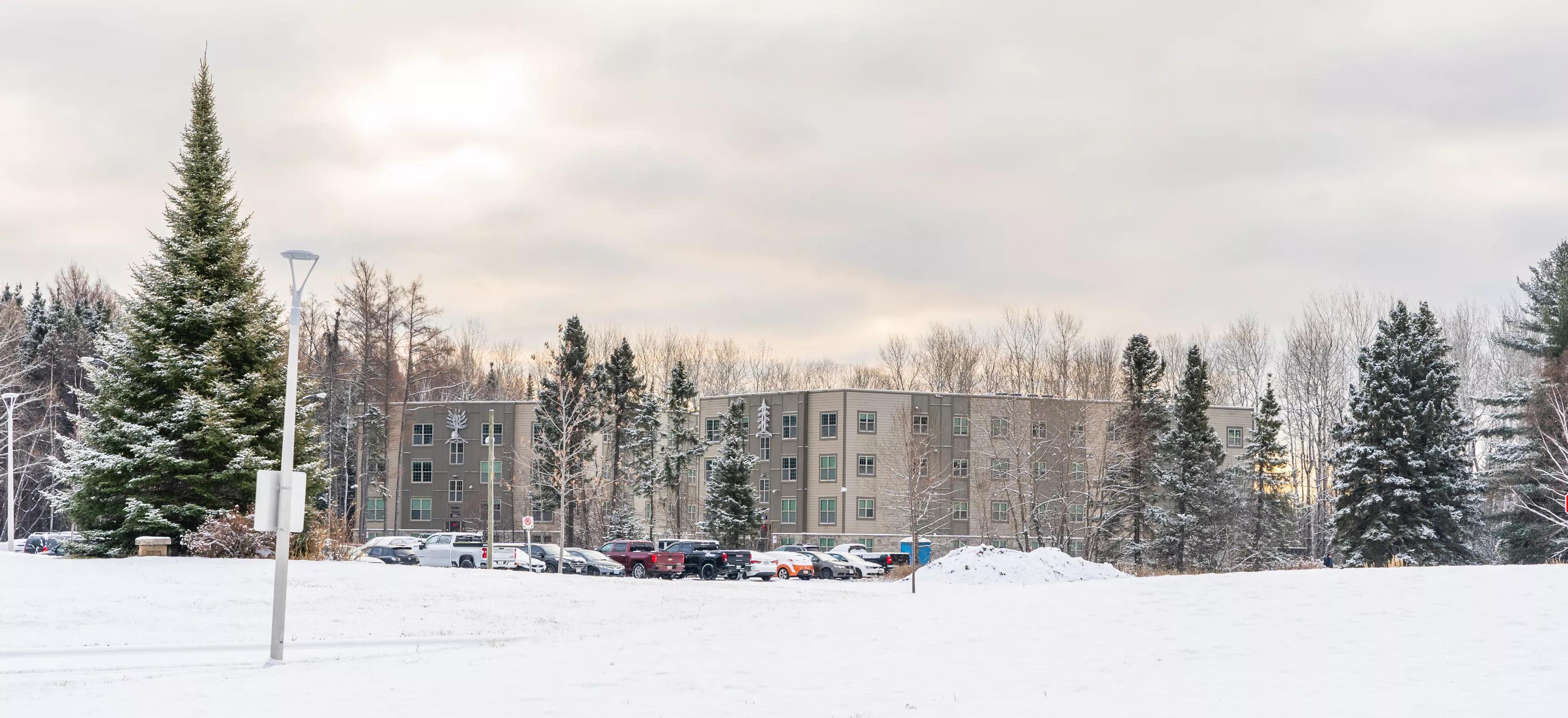 Snow-covered campus residence building with parked cars and winter trees