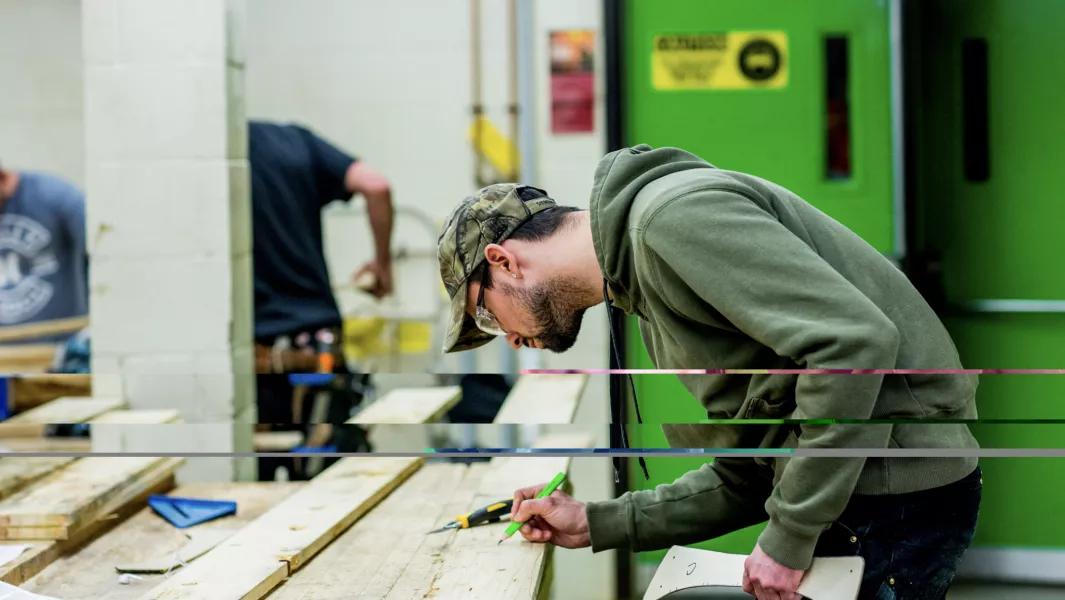 Carpentry student measuring and marking lumber at a workbench in a training shop