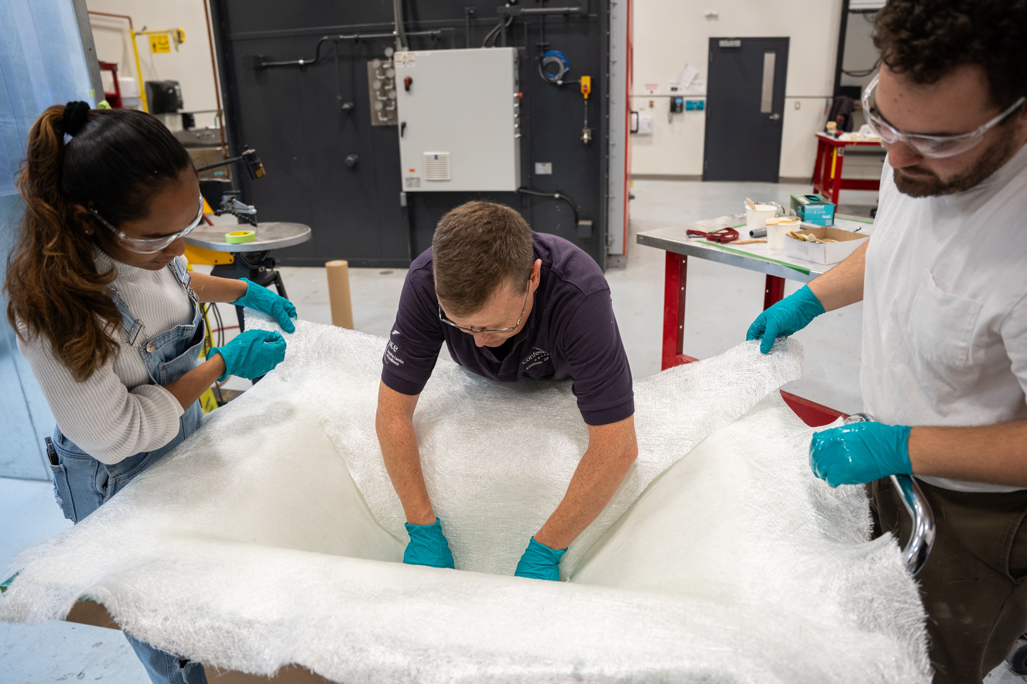 Three people in safety glasses and blue gloves working with a large white fibreglass sheet in a workshop