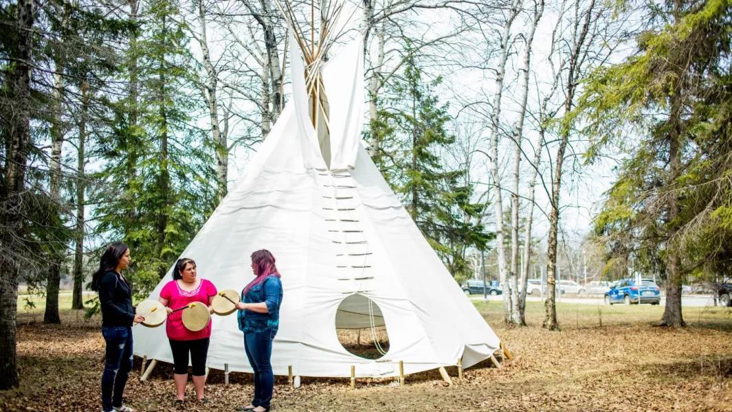 Group of women holding hand drums outside near a large tipi