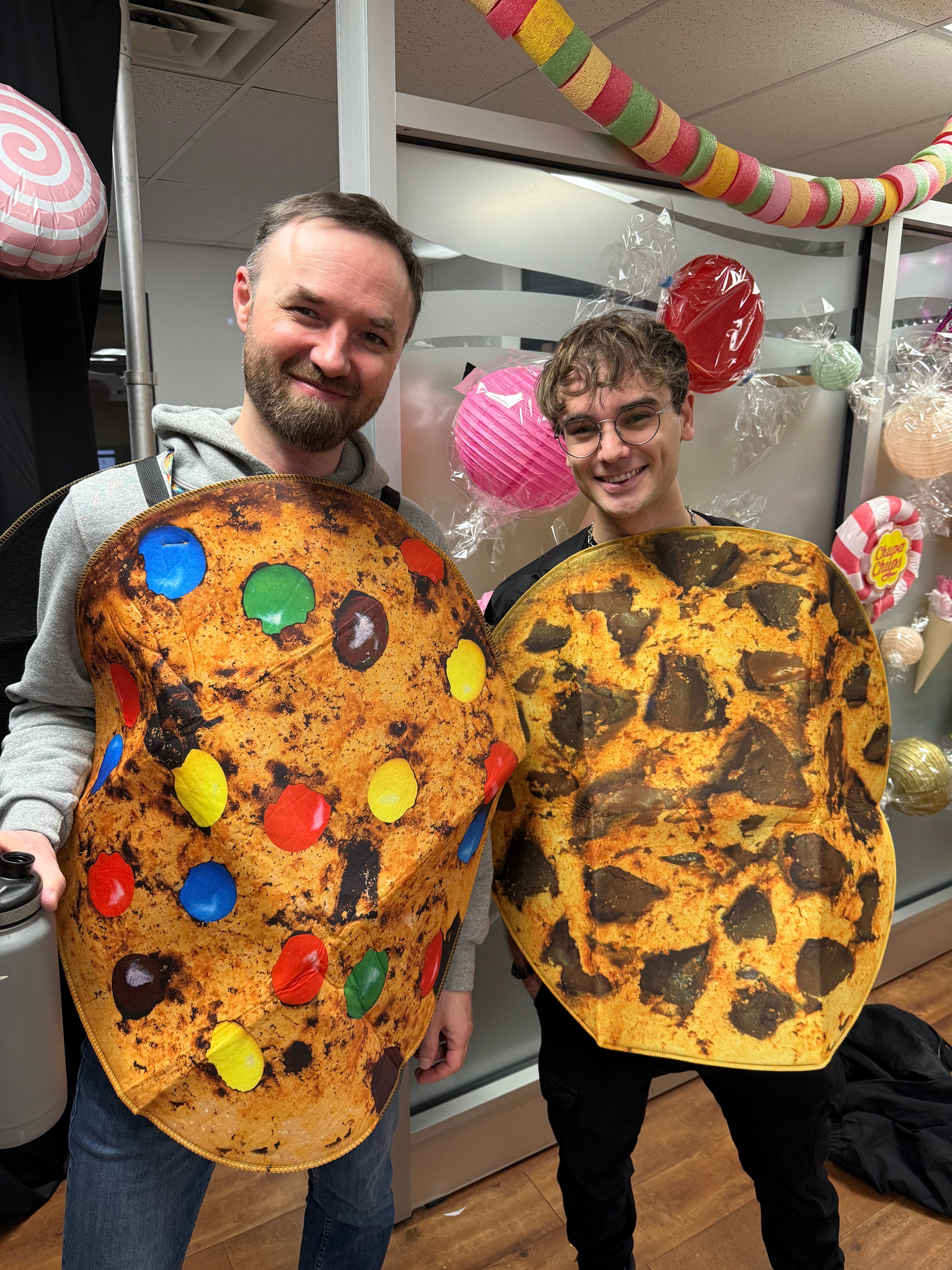 Two smiling people wear oversized chocolate chip cookie costumes in a decorated indoor space with colourful hanging decorations.