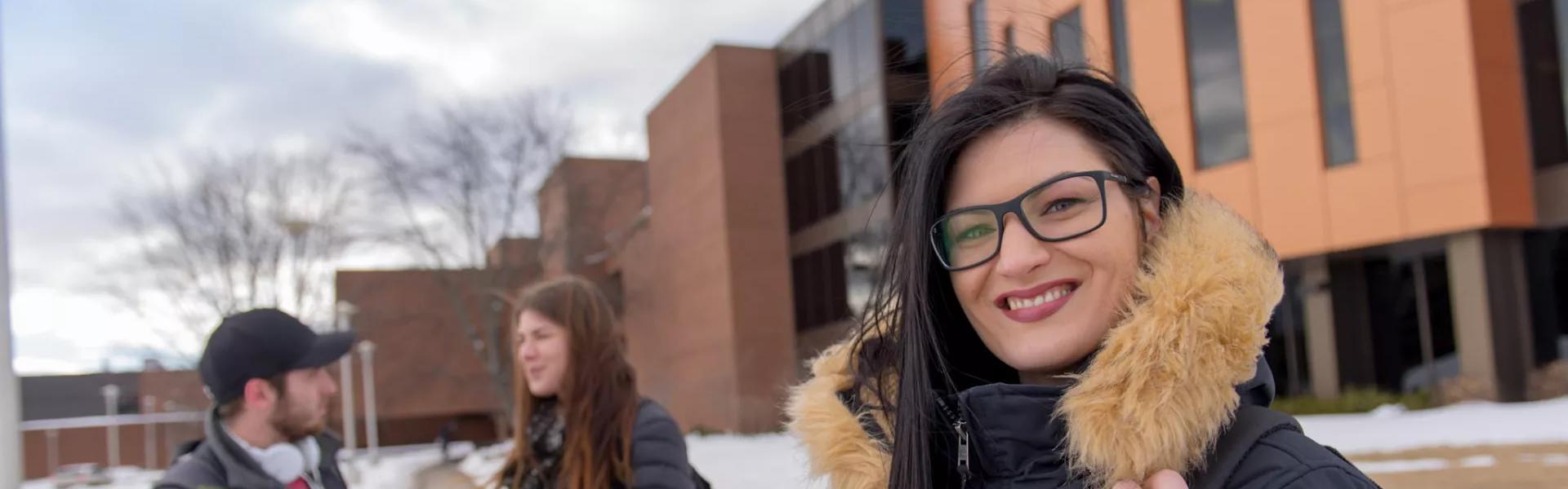 Student in glasses and winter coat smiling on snowy campus with classmates and buildings in background