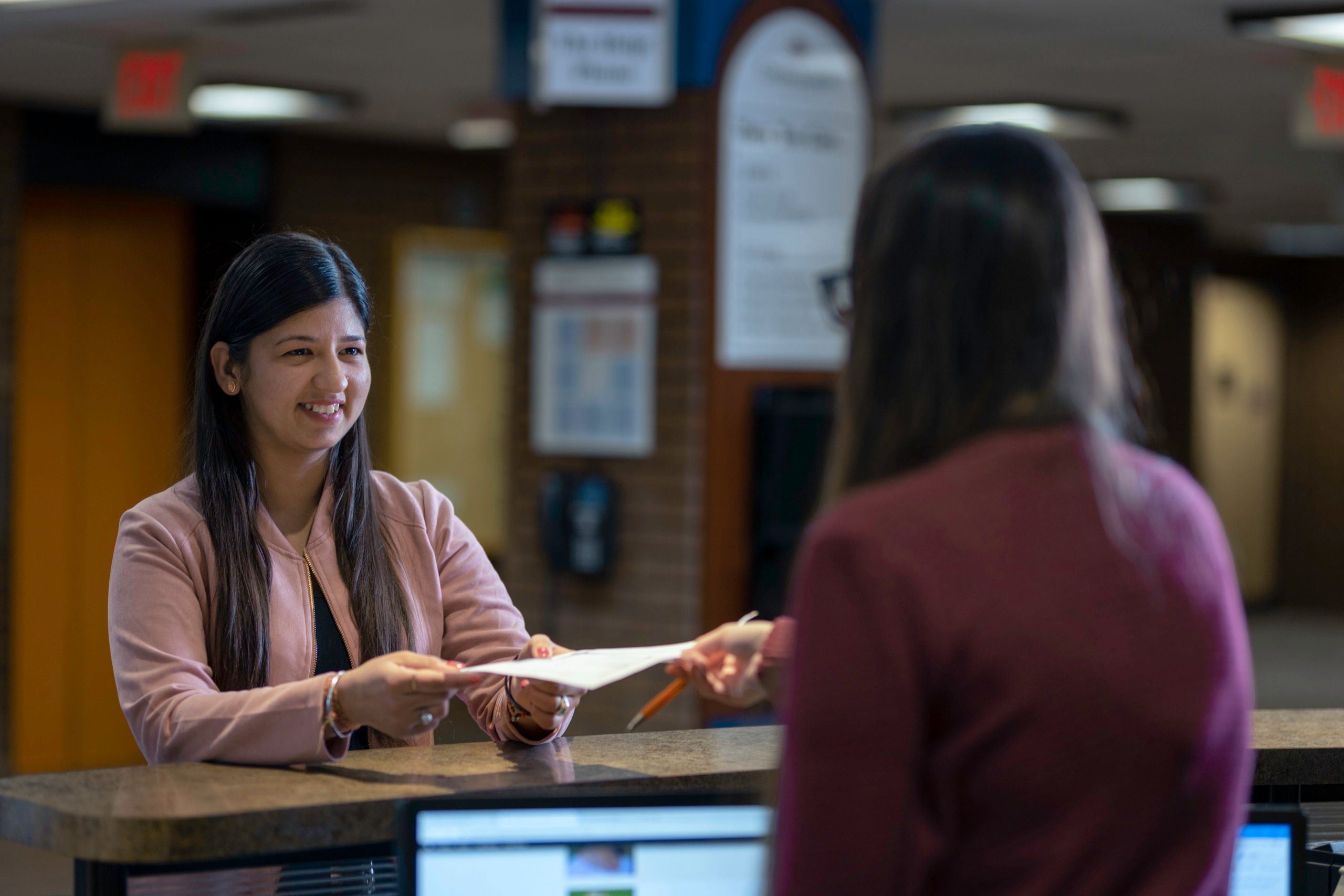A person at the library desk passes a sheet of paper to another person standing across the counter.