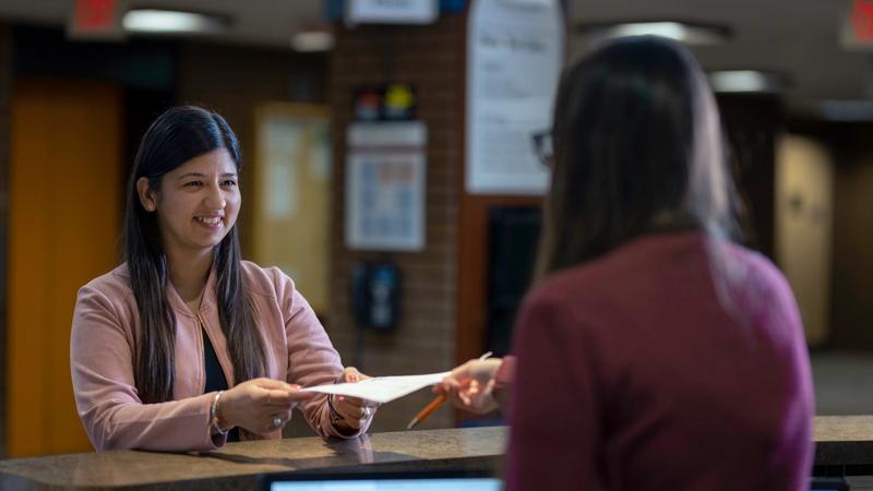 A person at the library desk passes a sheet of paper to another person standing across the counter.
