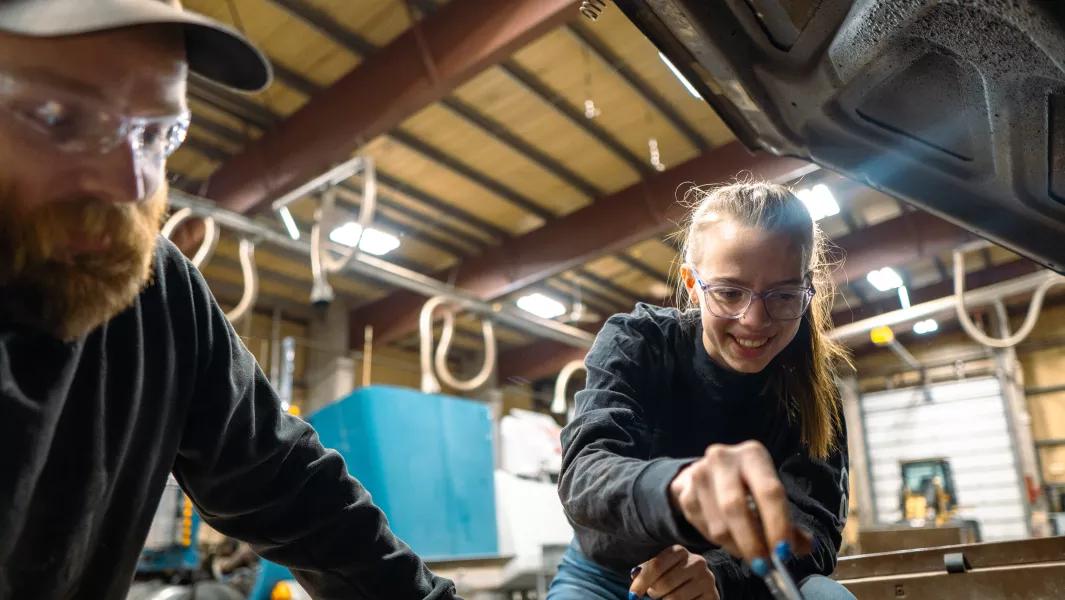 Student smiling while using a tool underneath a vehicle in a workshop