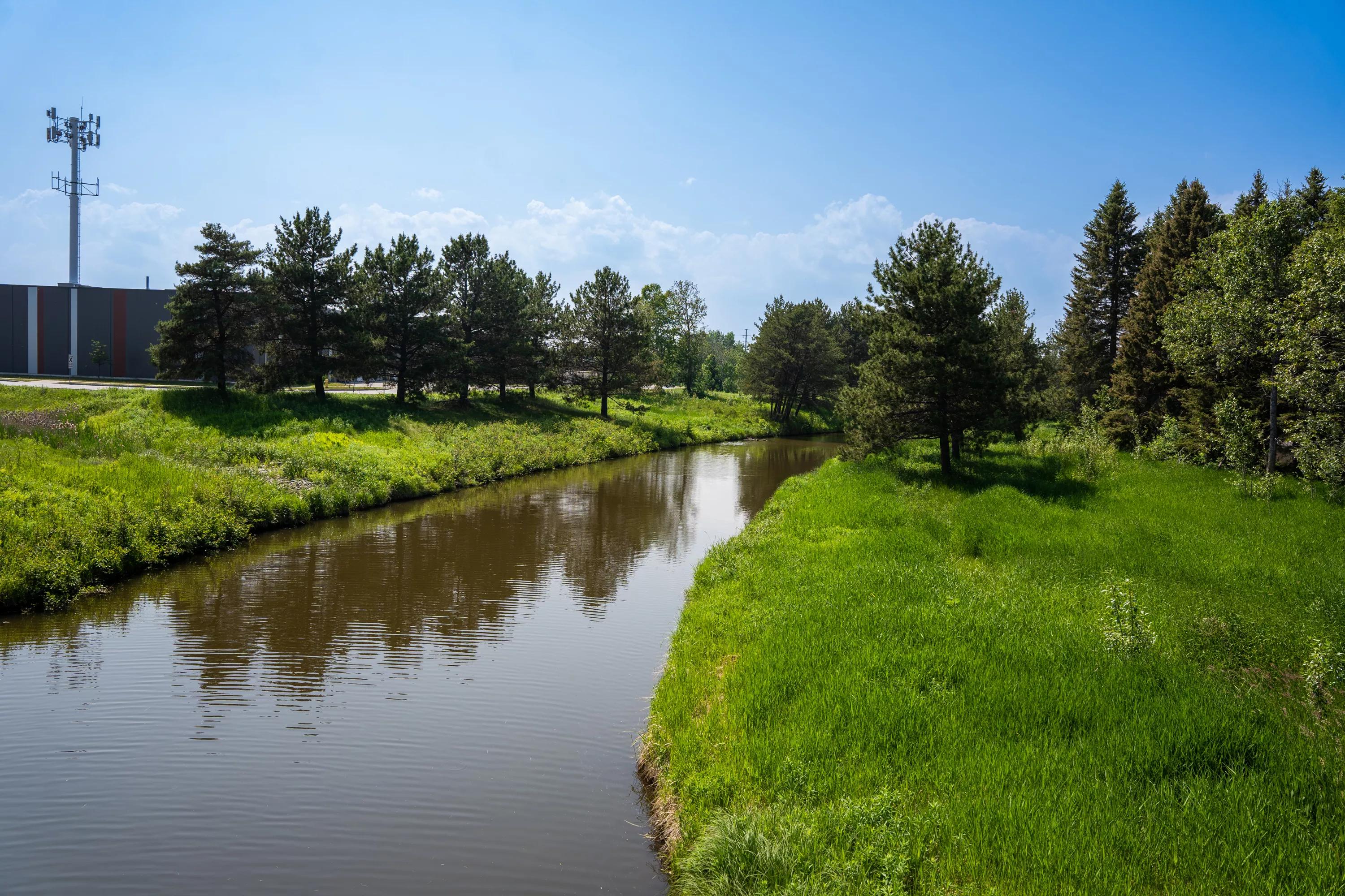 A narrow creek bordered by grass and trees under a clear blue sky