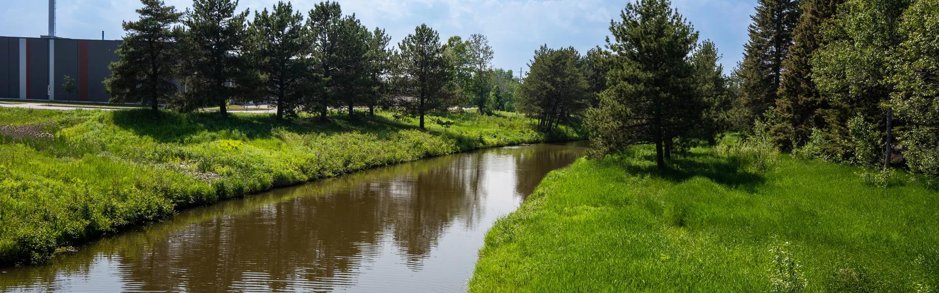 A narrow creek bordered by grass and trees under a clear blue sky