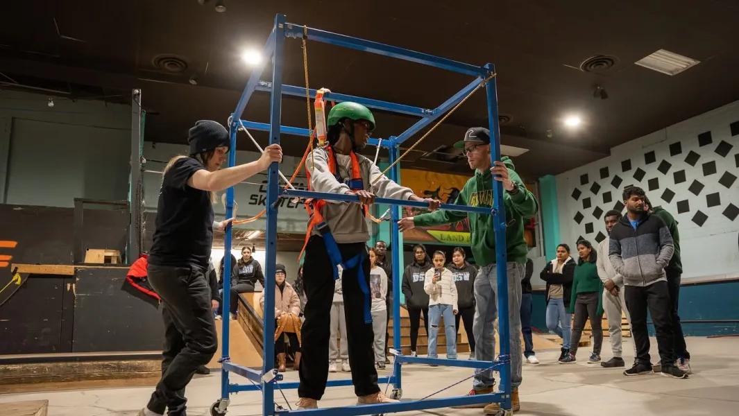 Students supporting a peer using a safety harness frame during an indoor activity