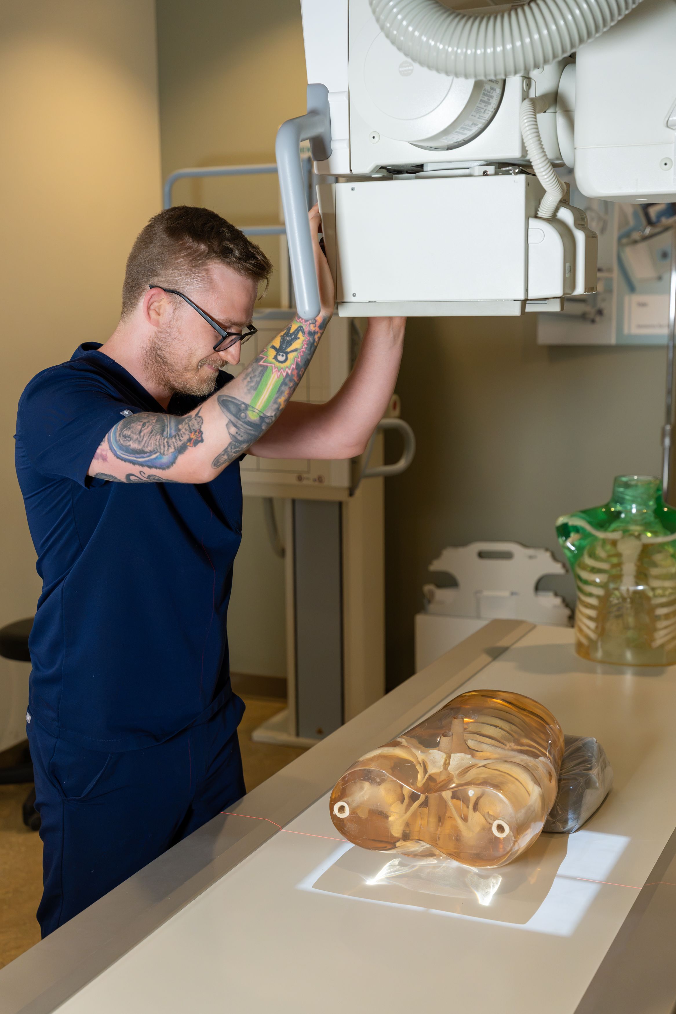 A person in navy scrubs adjusts an X-ray machine above a table holding a transparent anatomical model.