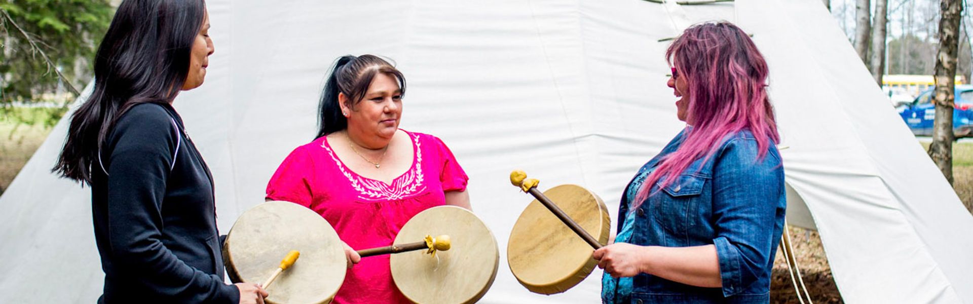 Three Indigenous women holding traditional hand drums outdoors in front of white tent structure