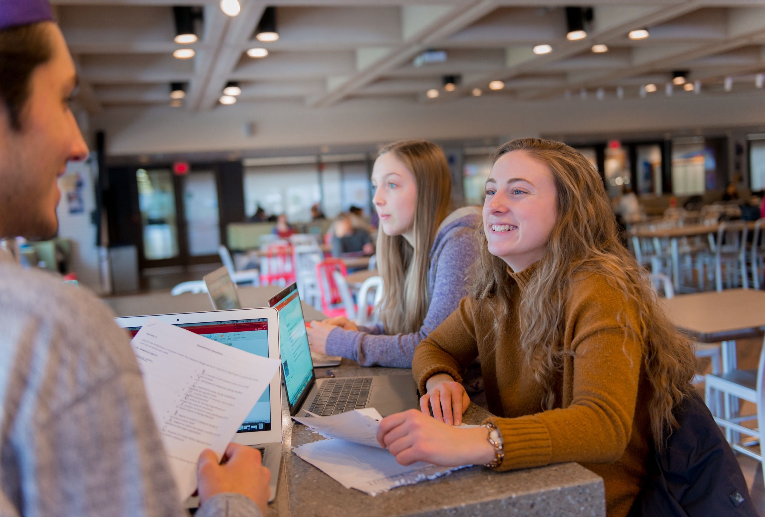 Two women sit at a library counter with laptops and papers, facing a person partly visible at the left.