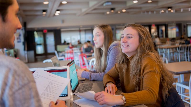 Two women sit at a library counter with laptops and papers, facing a person partly visible at the left.