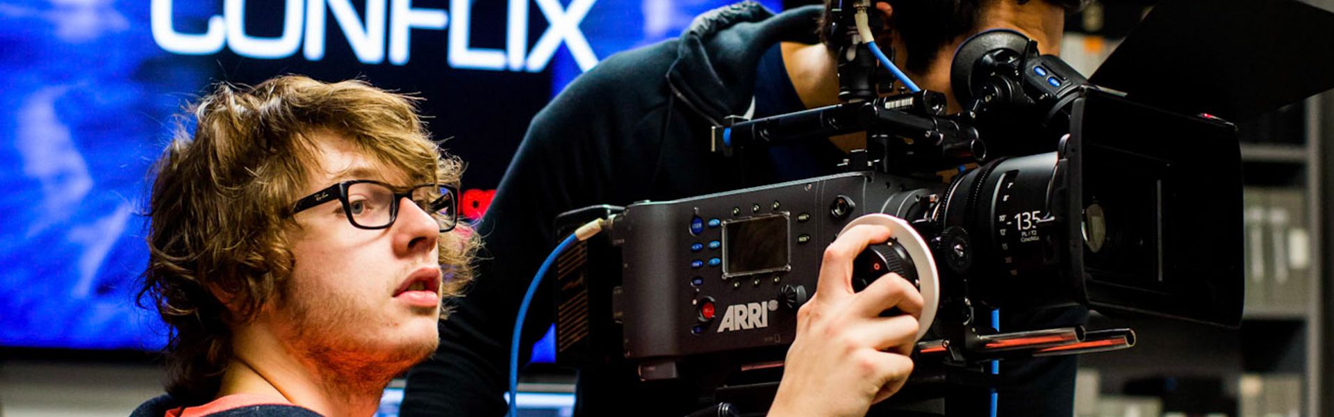 Student adjusts a professional video camera on a tripod in a studio with a “CONFLIX” screen in the background.
