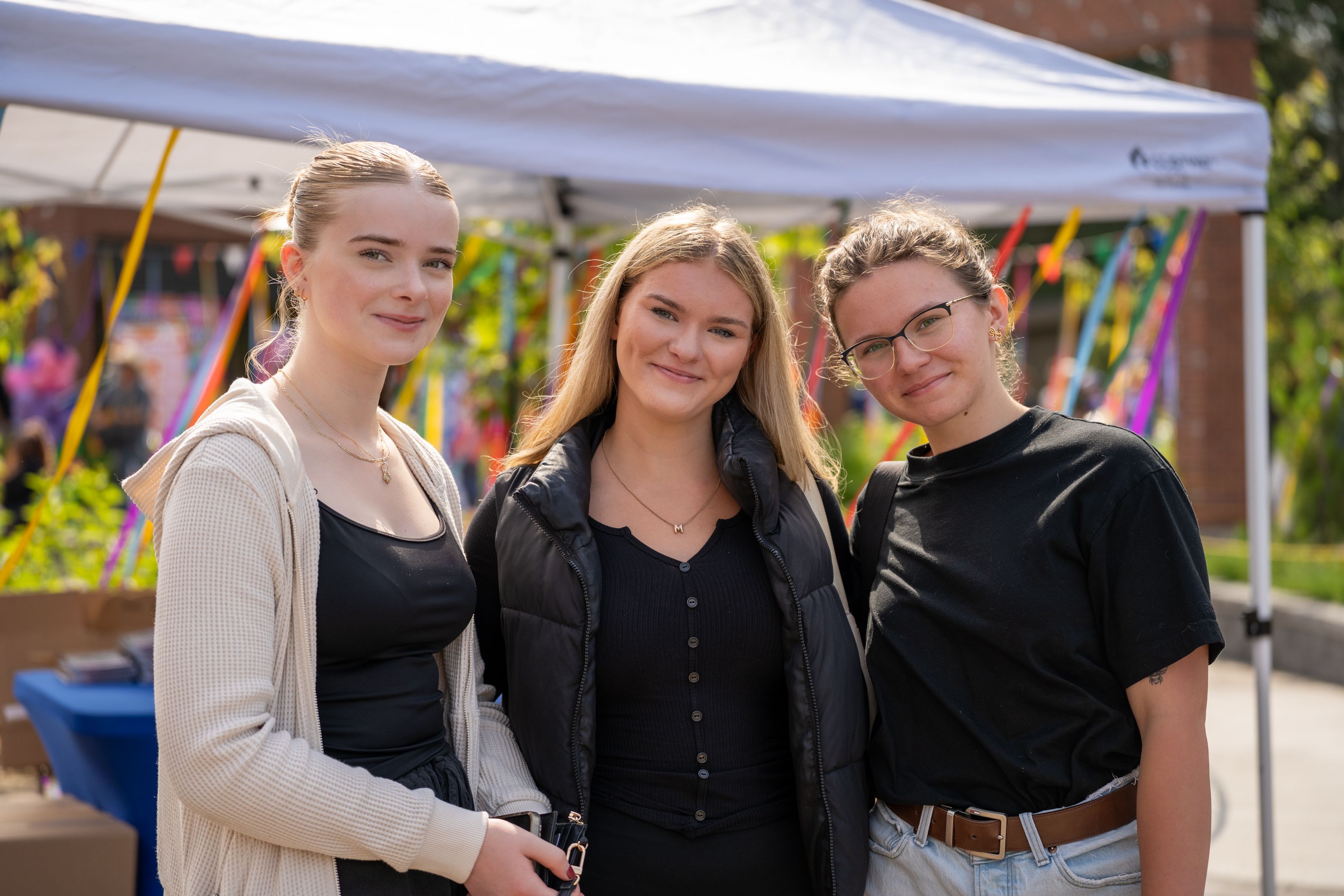 Three students standing outdoors under a tent, smiling. They are casually dressed and appear to be at a college event.