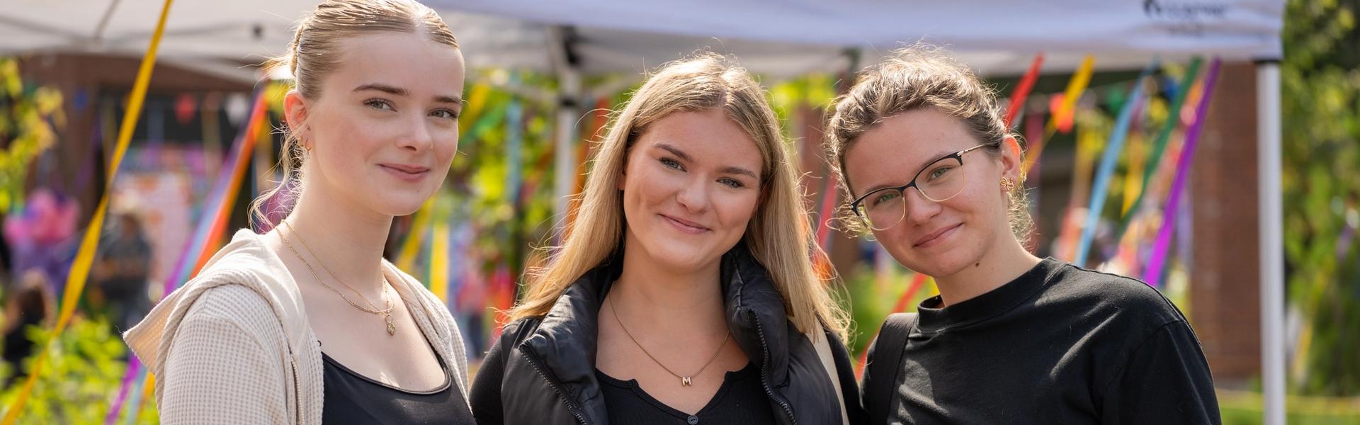 Three students standing outdoors under a tent, smiling. They are casually dressed and appear to be at a college event.