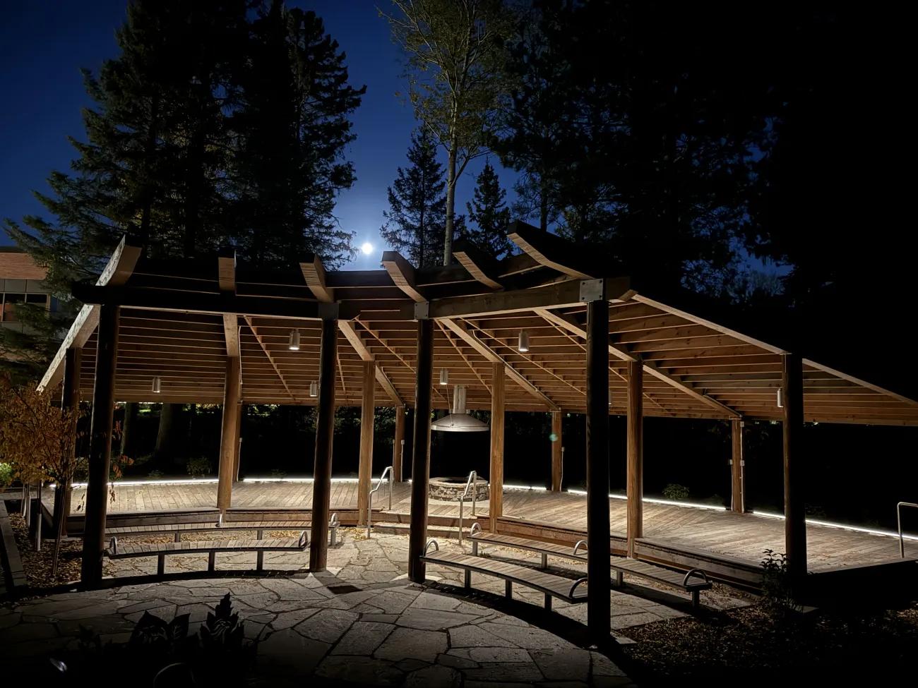 Illuminated wooden pavilion with seating at night, surrounded by trees on campus
