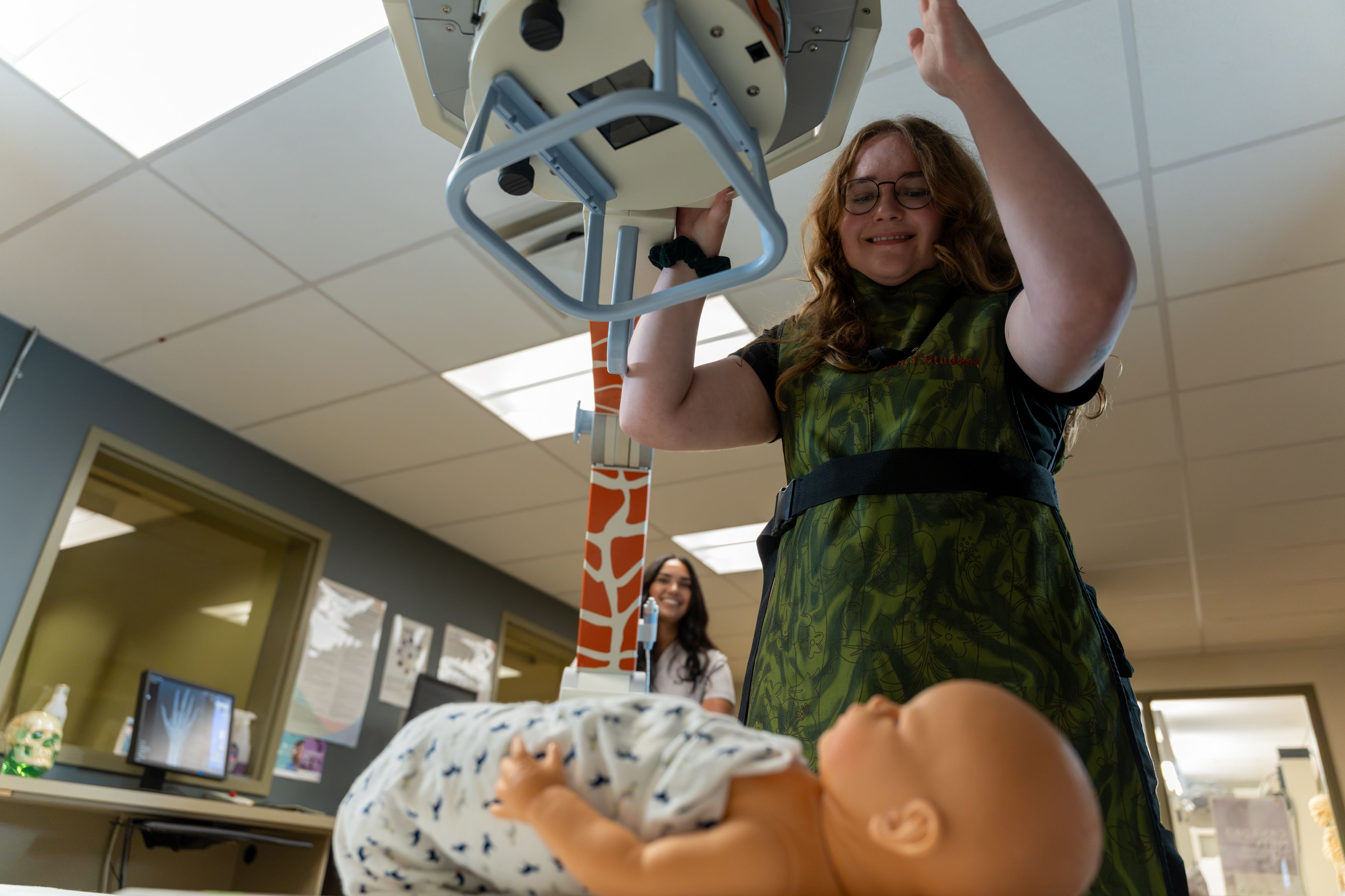 A student adjusts an overhead imaging machine above a baby mannequin in a classroom, while another person watches.