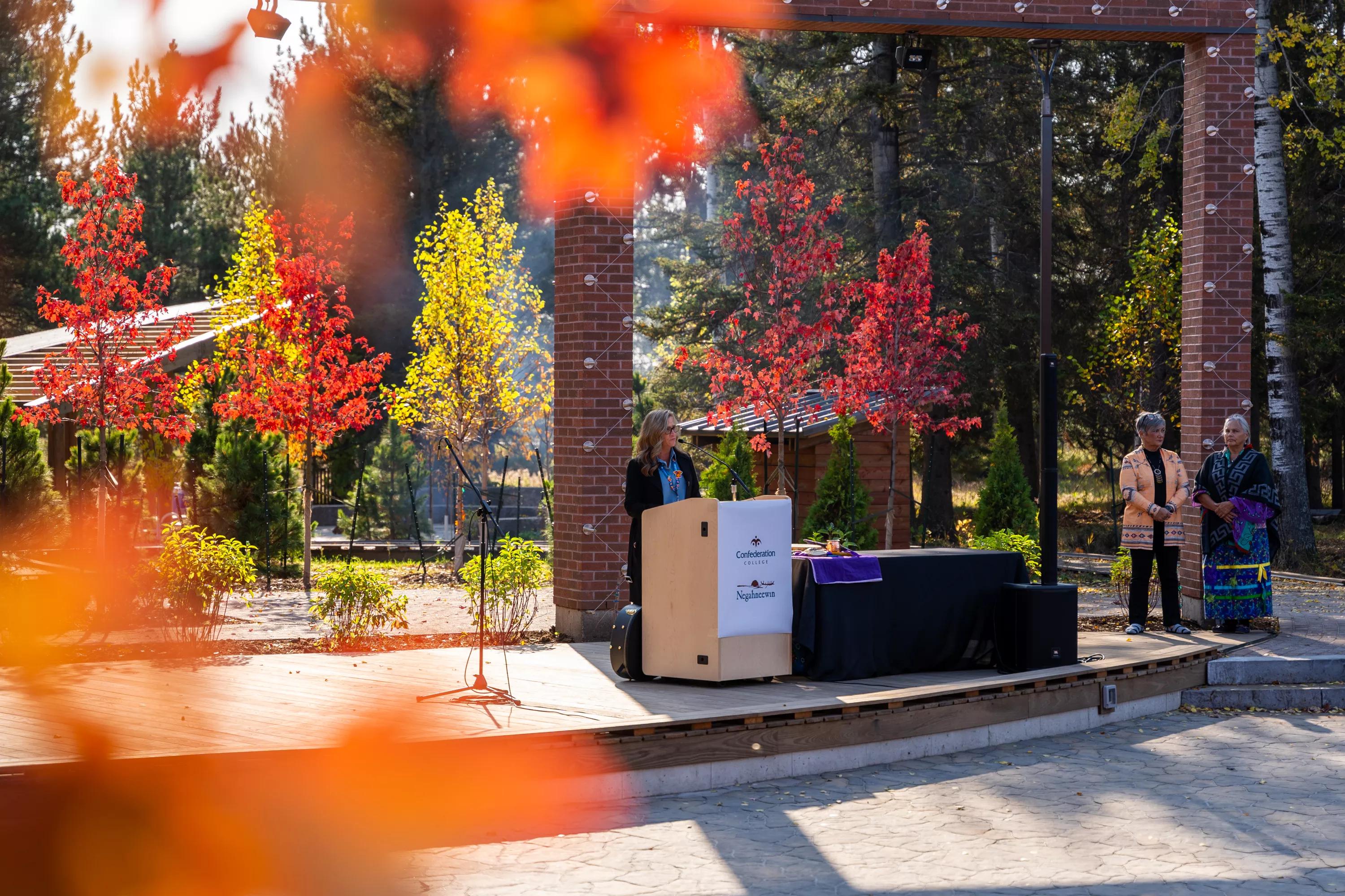 Speaker at podium during outdoor campus ceremony, framed by autumn trees, with two individuals standing nearby.
