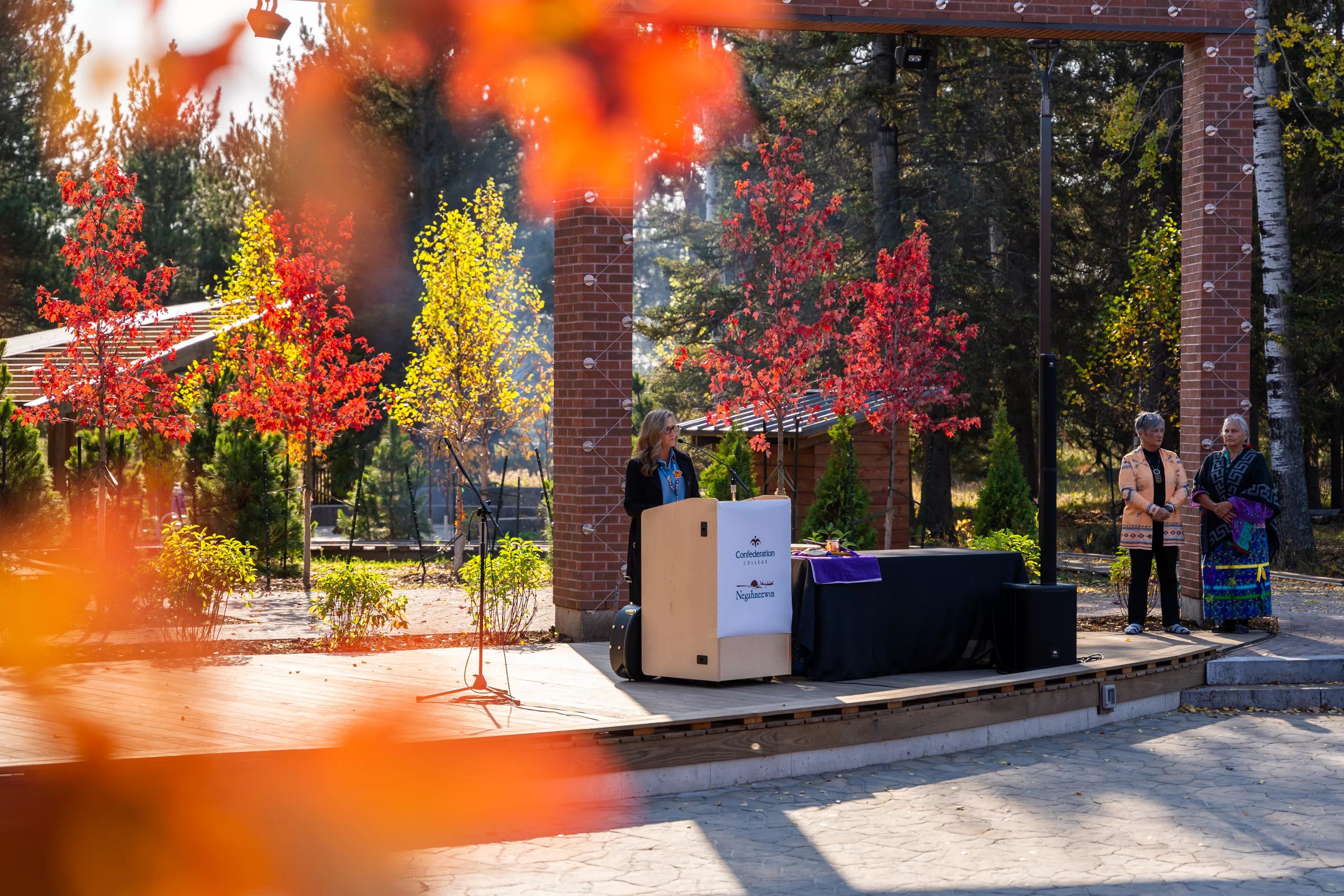 Speaker at podium during outdoor campus ceremony, framed by autumn trees, with two individuals standing nearby.