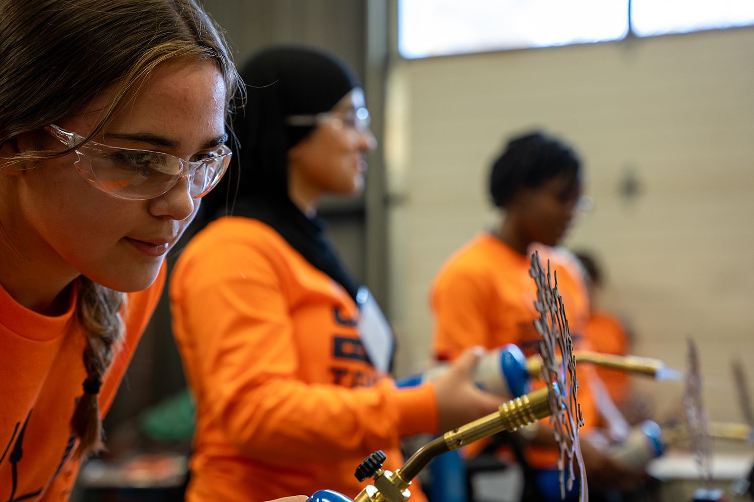 People in orange shirts and safety glasses work with metal pieces and a torch at a trades workshop.