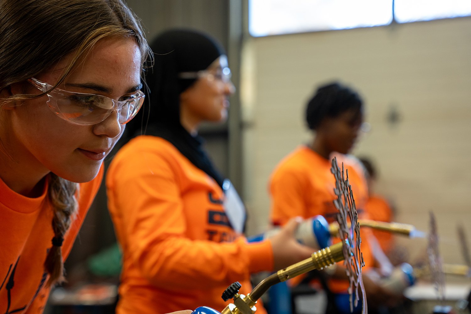People in orange shirts and safety glasses work with metal pieces and a torch at a trades workshop.