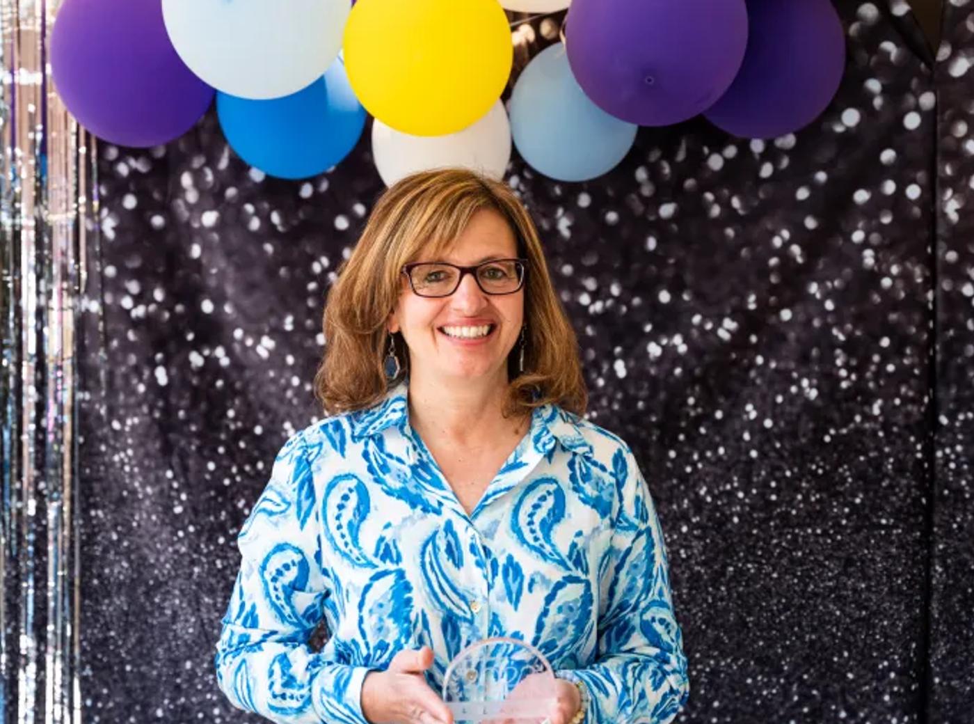 A person holding an award, smiling, in front of a balloon and glitter backdrop