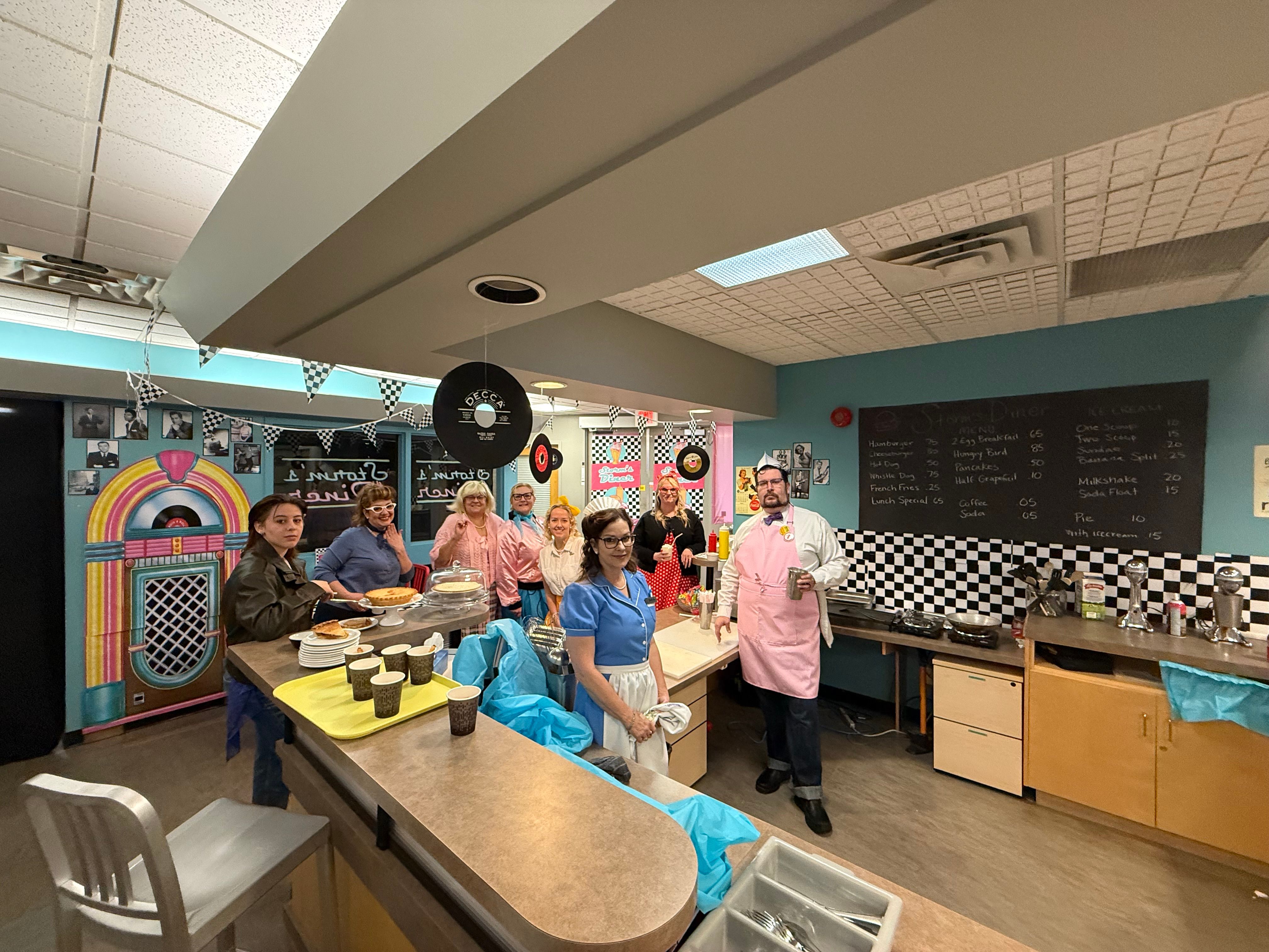 A group in colourful costumes stand behind a café counter decorated with checkered patterns and record-themed décor.