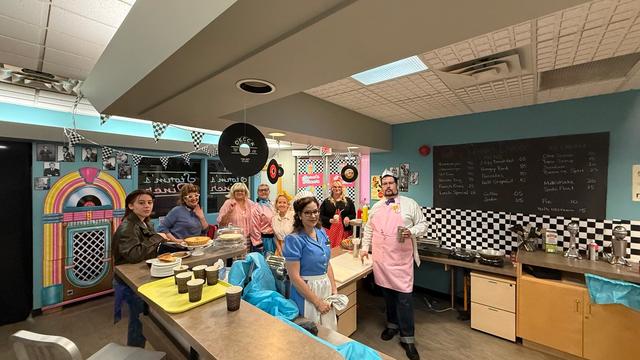 A group in colourful costumes stand behind a café counter decorated with checkered patterns and record-themed décor.