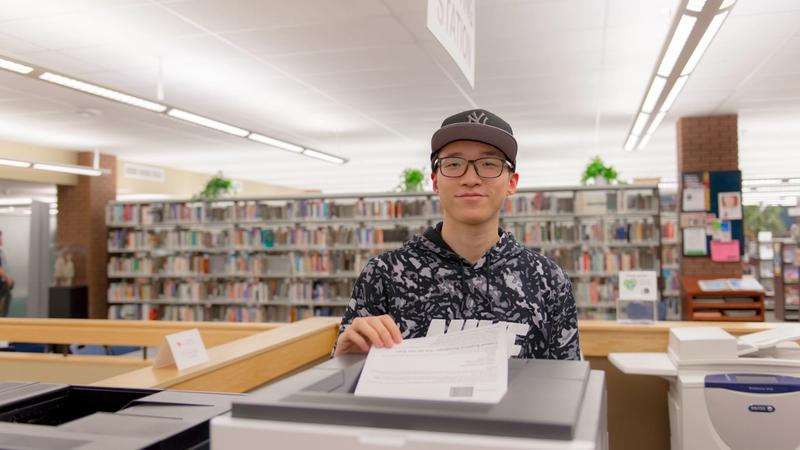 A person in glasses and a patterned hoodie holds a sheet of paper beside a library printer, with bookshelves behind.