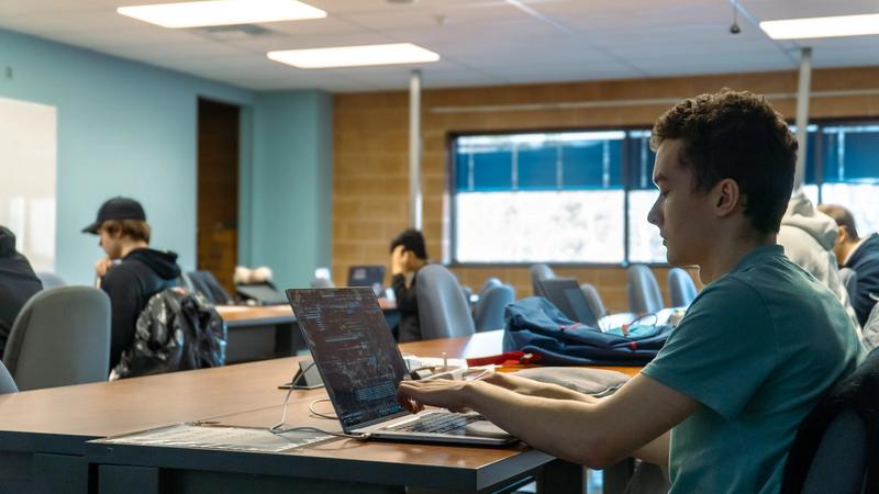 Students working at laptops in a classroom with tables, chairs, and large windows