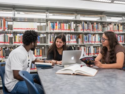 Three students sitting at a table in a library surrounded by bookshelves, with open books and a laptop