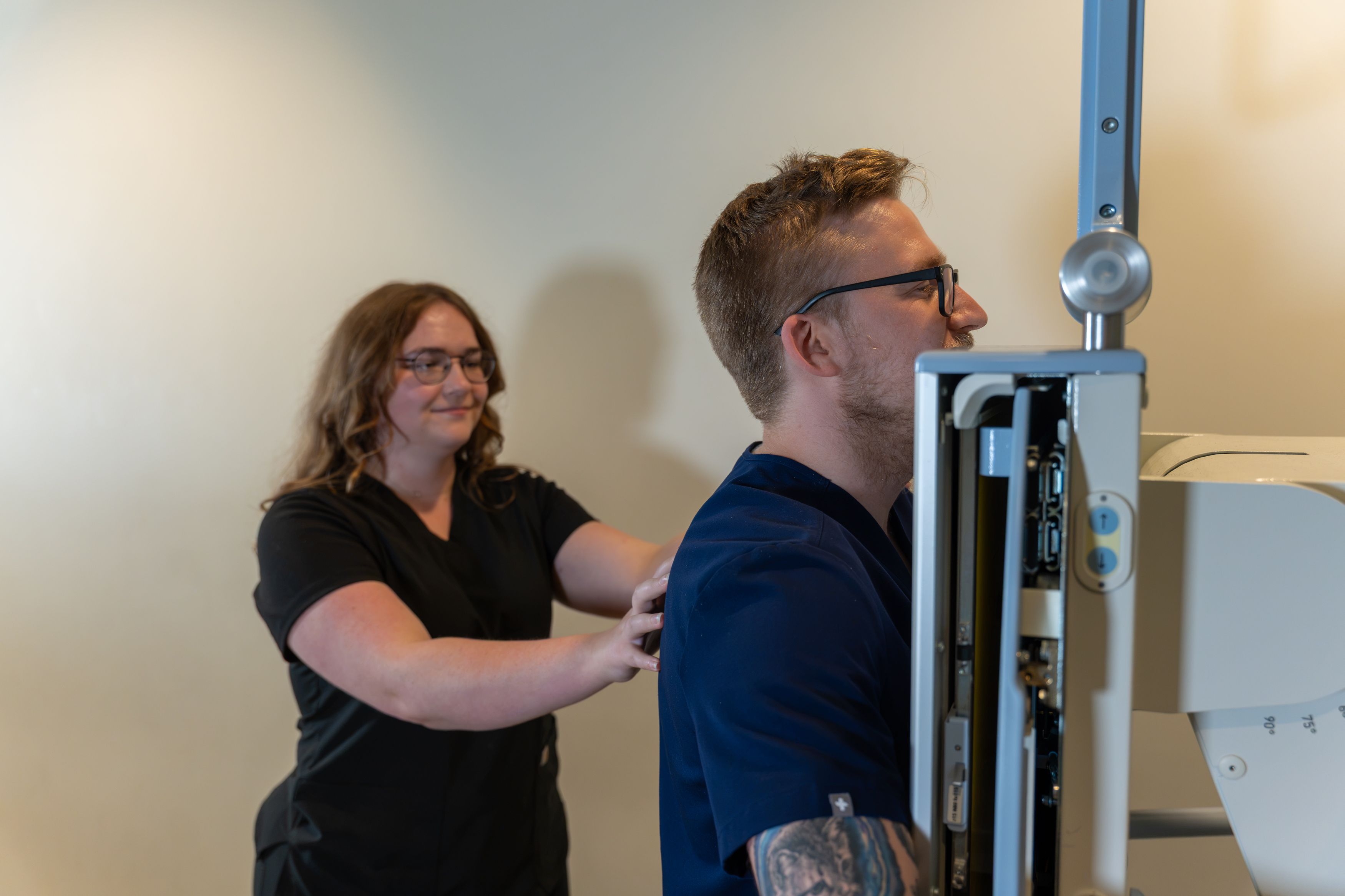 A woman adjusts a man in glasses and a blue shirt beside a medical imaging machine in a clinical room.