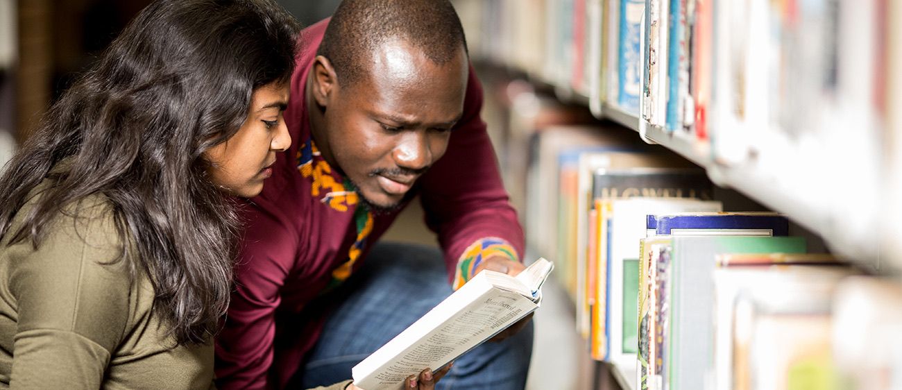Two people crouch beside library shelves, looking together at an open book held between them.