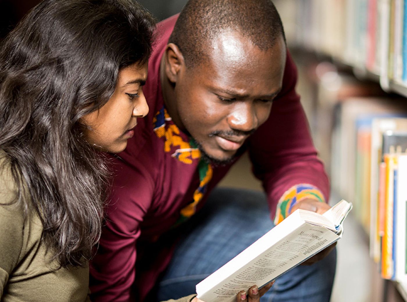 Two people crouch beside library shelves, looking together at an open book held between them.