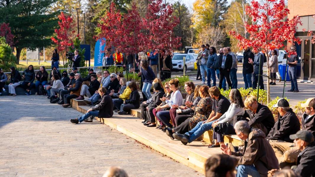 Audience seated outdoors at a campus gathering among autumn trees