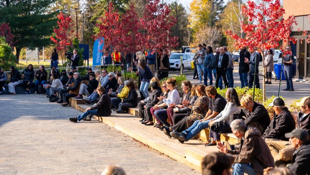 Audience seated outdoors at a campus gathering among autumn trees