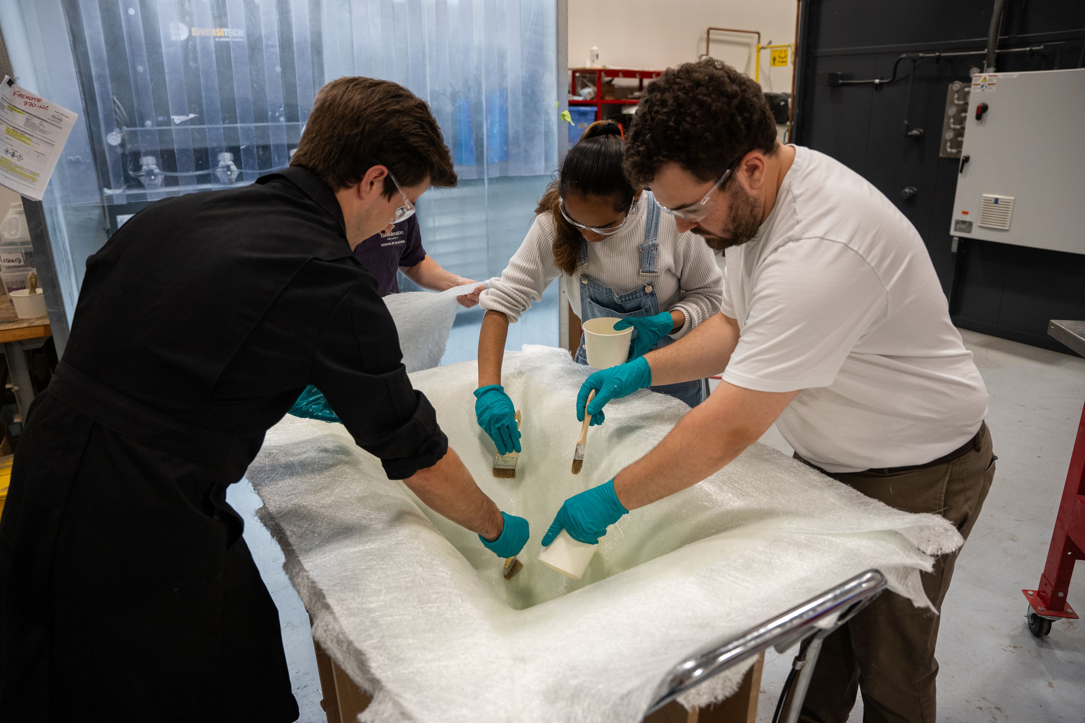 Three people wearing safety glasses and gloves brush resin onto a large white fabric piece in a workshop
